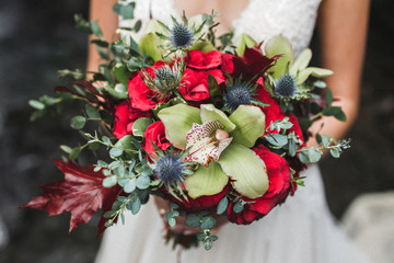 Bride holding in hands elegant beautiful bouquet with red roses, yellow orchid and green leaves close-up