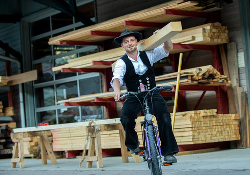 Zimmermann mit Balken auf der Schulter fährt Fahrrad vor Holzlager