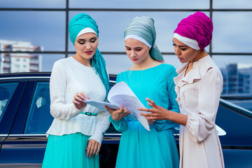 group of three successful business multinational Muslims women in stylish veiled hijab a turban headscarf is tied up on the head communicating and talking about work office in street skyscraper view