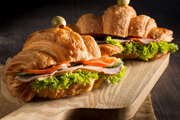 Photo of tasty and fresh croissant sandwich with salad, ham, cheese, tomatoes on wooden background. Morning breakfast concept. Healthy and fast food.