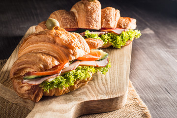 Photo of tasty and fresh croissant sandwich with salad, ham, cheese, tomatoes on wooden background. Morning breakfast concept. Healthy and fast food.