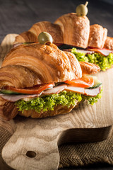 Photo of tasty and fresh croissant sandwich with salad, ham, cheese, tomatoes on wooden background. Morning breakfast concept. Healthy and fast food.