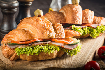 Photo of tasty and fresh croissant sandwich with salad, ham, cheese, tomatoes on wooden background. Morning breakfast concept. Healthy and fast food.