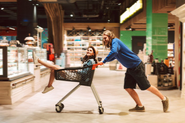 Young guy pushing shopping trolley with pretty girl inside while happily spending time together in modern supermarket