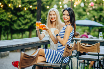 Young blond and brunette women friends with red lips holding iced aperol spritz in modern outdoor...