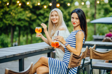 Young blond and brunette women friends with red lips holding iced aperol spritz in modern outdoor cafe on the green park. Wearing in striped and white dress.