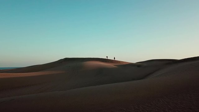 Silhouette of two tourists crossing sand dune in a desert in sunset light with an ocean on the background. Beautiful oringe desert landscape. Two travelers on adventure in a desert. Gran Canaria