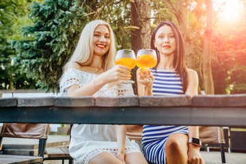 Blond and brunette sweden women drinking aperol spritz, clink glasses, looking at camera in modern outdoor cafe on the green park. Wearing in striped and white dress. Copy space