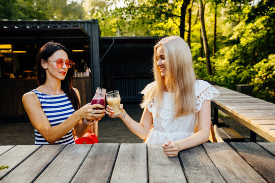 Close up portrait of bright summer portrait of happy sister girls having great time at green outdoor cafe in park, hipster best friends making cheers with tasty cocktails.