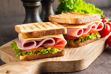 Close-up of two sandwiches with bacon, salami, prosciutto and fresh vegetables on rustic wooden cutting board. Club sandwich concept.