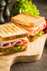 Close-up of two sandwiches with bacon, salami, prosciutto and fresh vegetables on rustic wooden cutting board. Club sandwich concept.