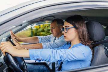  Young serious inexperienced woman driving a car in alert, worried instructor man sitting aside and...