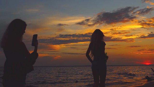 A young girl enjoys the view of the sea on the beach and takes pictures on the phone other girl. Silhouetted woman at sunset beach. Silhouettes at sunset.