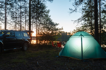 Night landscape with a tent in the forest near lake. The light from the lantern in a tent. Camping in the wild nature. Car and portable table and chairs, green tourist tent.  