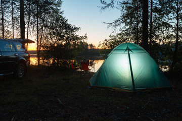 Night landscape with a tent in the forest near lake. The light from the lantern in a tent. Camping in the wild nature. Car and portable table and chairs, green tourist tent. 