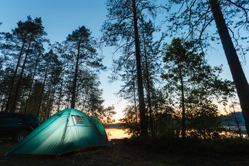 Night landscape with a tent in the forest near lake. The light from the lantern in a tent. Car and portable table and chairs, green tourist tent. Romantic evening with a tent at night.