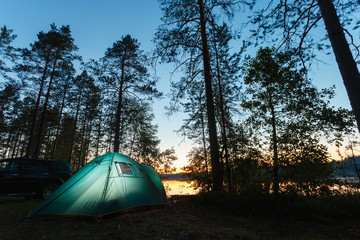 Night landscape with a tent in the forest near lake. The light from the lantern in a tent. Car and portable table and chairs, green tourist tent. Romantic evening with a tent at night.