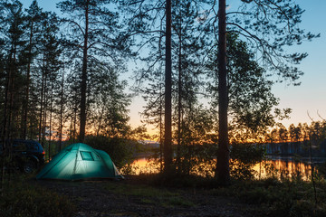 Night landscape with a tent in the forest near lake. The light from the lantern in a tent. Car and portable table and chairs, green tourist tent. Romantic evening with a tent at night.