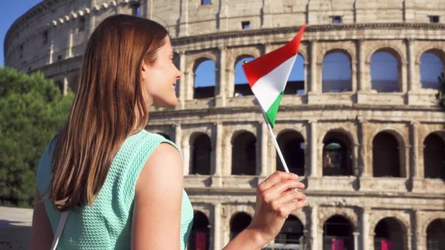 Young woman standing near famous attraction Colosseum in Rome, Italy. Teenage girl waving Italian flag in slow motion. Happy female tourist enjoying european vacation. Student travel through Europe