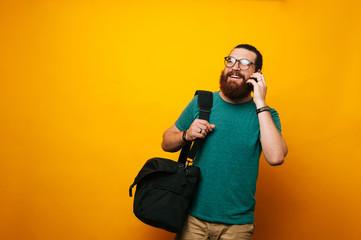 Cheerful young bearded man talking on the phone going to work with his shoulder bag.