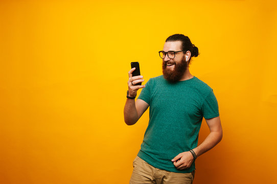 Young smiling bearded man using phone wearing earpods and glasses is taking a selfie.