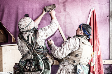 Marines in combat uniform, body armor and ammo belts hammering nail into wall with sledgehammer to hang flag of United States Marine Corps in temporary camp. Arranging military outpost on front line