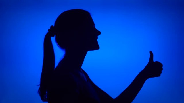 Silhouette of young woman making thumb up gesture on blue background. Female's face in profile showing thumbs-up sign. Black contur shadow of teenager's half-face. Success and achievement concept