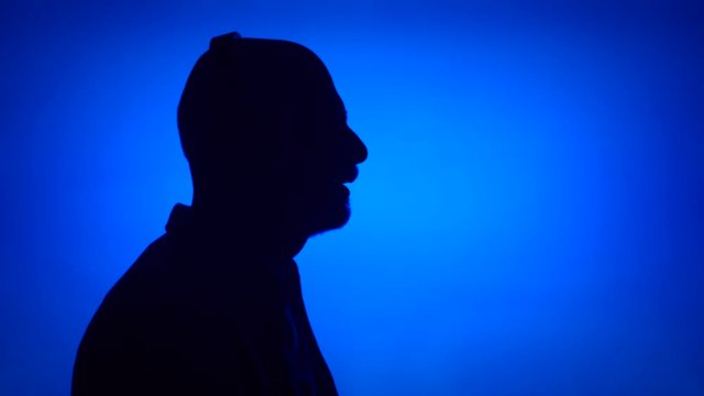 Silhouette of senior man remove big black wireless headphones on blue background. Male's face in profile listening to music. Black contur shadow of grandfather's half-face singing