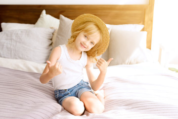 Obraz na płótnie Canvas beautiful baby girl blonde in hat sitting on bed on Sunny Sunday day. summer vacation. thumbs up.