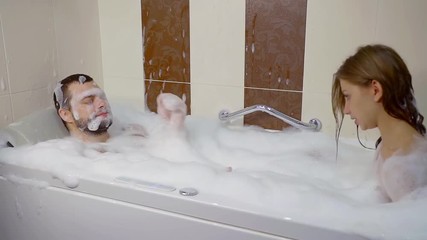 young honeymooners are taking a bath together, man is relaxing in foam and woman talking with him