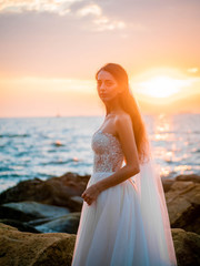 Portrait of girl in wedding luxury dress watching sunrise on sea shore. Bride on a rocks. Beautiful waves near to her. Woman enjoying happy moments with nature.