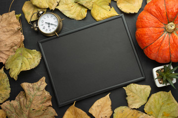an alarm clock, a pumpkin and autumn foliage and an empty black frame. flat lay season food items top view