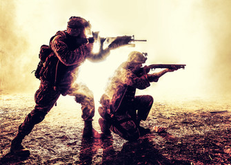 Silhouettes of two army soldiers, U.S. marines team in action, surrounded fire and smoke, shooting...