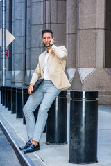 European Businessman traveling, working in New York City, with beard, little gray hair, wearing beige blazer, gray pants, leather shoes, holding briefcase, sitting on street, talking on cell phone..