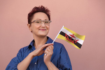 Brunei flag. Woman holding  Brunei flag. Nice portrait of middle aged lady 40 50 years old with a national flag over pink wall background outdoors.