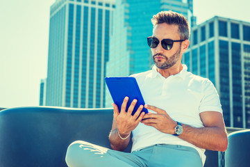 Modern Reading. Young European Man with beard, little gray hair, wearing white Polo shirt, sunglasses, sitting in business district with high buildings in New York City, reading blue tablet computer.. - Powered by Adobe
