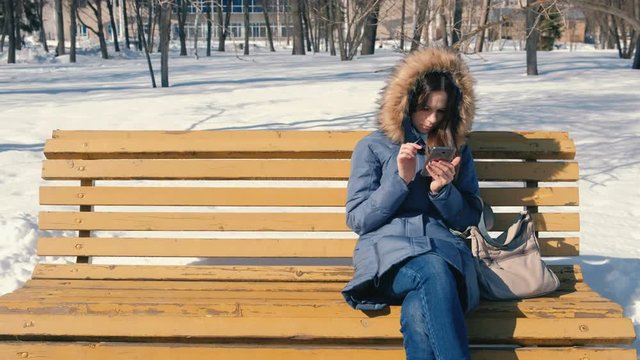 Woman is browsing internet on her phone sitting on the bench in winter city park in sunny day.