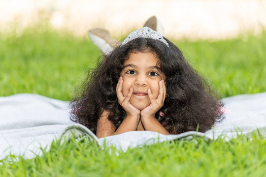 A young girl surrounded by green  is laying on a white blanket
