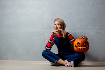 Portrait of a young style blonde girl in red striped sweater with halloween pumpkin on grey background.
