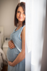 Beautiful brunette woman in a 7th month pregnancy in blue dress grabs her tummy next to bright window. light interior bedroom