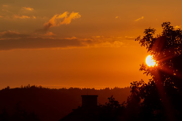 Sunrise with sun partially covered by a tree and with the silhouette of a house and orange cloudy sky