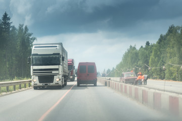 A lorry and truck on the road with concrete white and red blocks from one side and safety rail or barrier from other side. Part of the highway is under construction