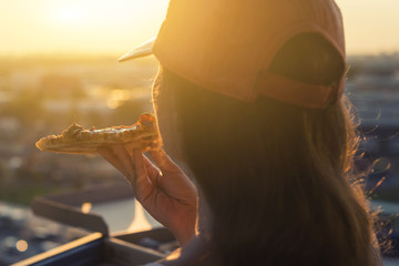 A female in a sports suit holds a huge piece of pizza on sunset and city background.