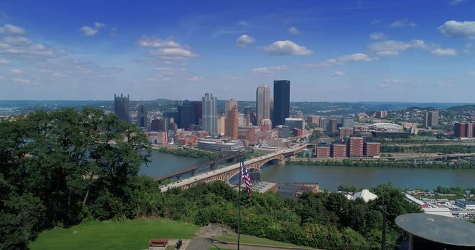 A wide aerial establishing shot of the iconic Pittsburgh, Pennsylvania skyline on a summer day as seen from Emerald View Park. American flag in the foreground.  	