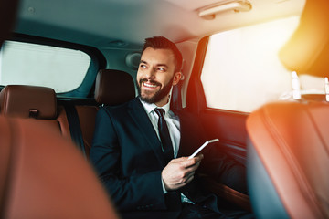 Handsome businessman with phone on hand sitting on the backseat of the car, guy looking away