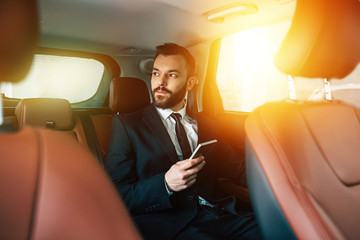 Confident bearded Businessman using mobile phone on back seat in car and looking away while trip