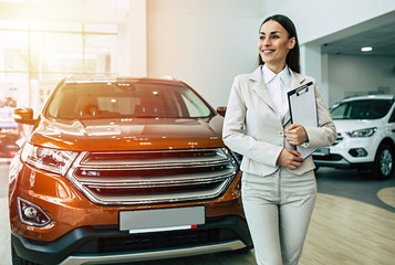 Beautiful smiling saleswoman in full suit in dealership on cars background with documents in tablet...