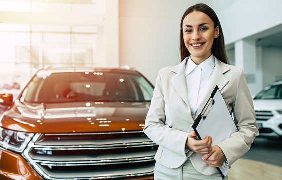 Beautiful smiling saleswoman in full suit in dealership on cars background with documents in tablet in hands