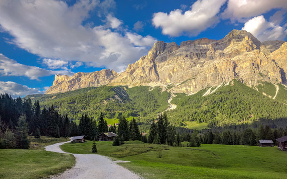 Green meadow with a gravel path, sheds and trees with high mountains and cloudy blue sky in the background