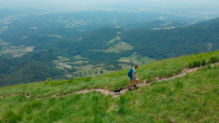 Fototapeta premium AERIAL: Athletic girl and her lovely dog walk up a steep mountain trail in Alps